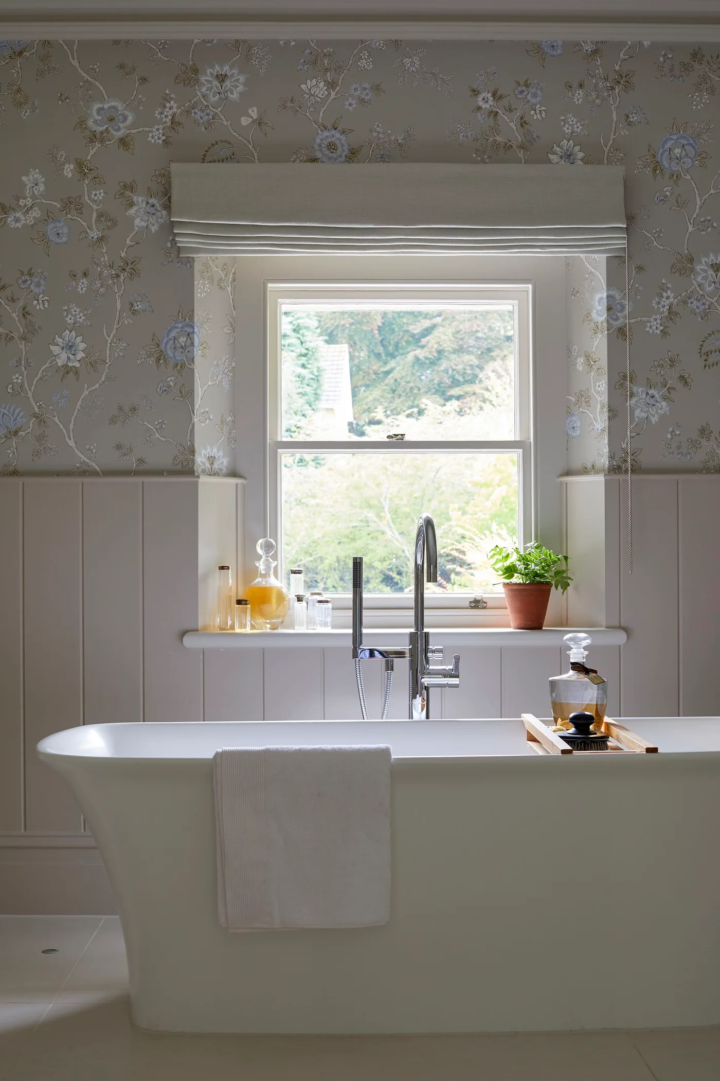 Bathroom with pale vertical paneling, floral wallpaper around a window, a fitted blind and a white freestanding bath.