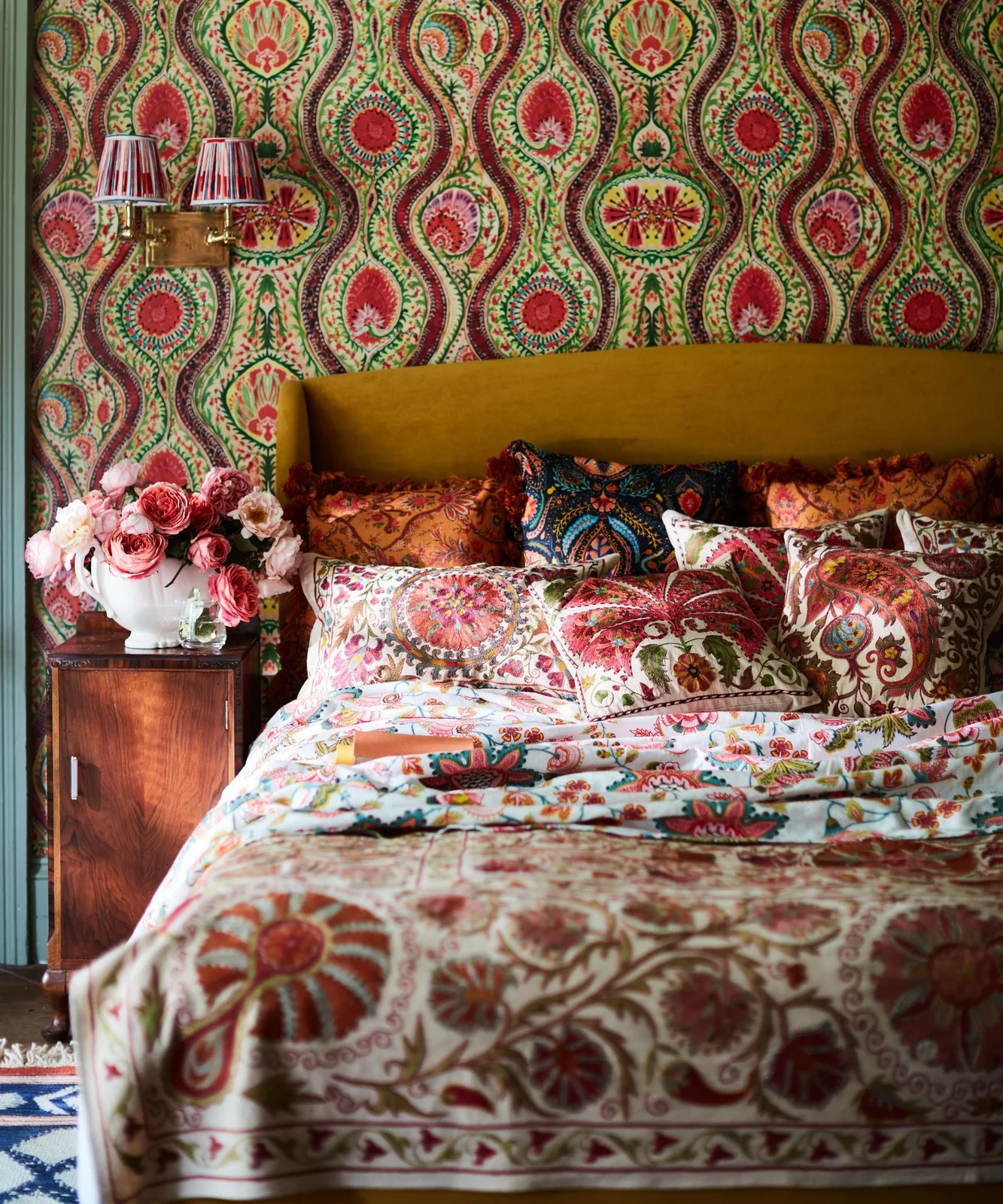 Red-and-green paisley wallpaper rises behind a mustard headboard, multicolored bedding and striped wall-light shades