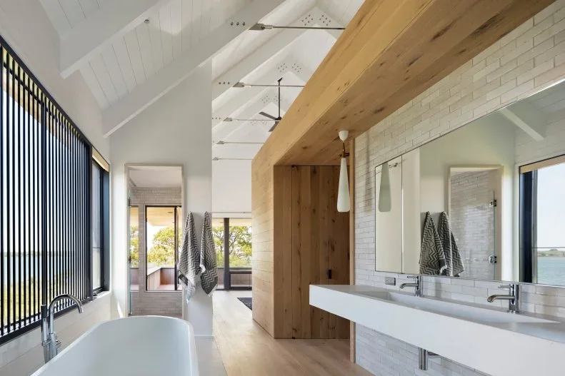 A long white basin and broad mirror sit beneath a wood beam-like feature beside white brickwork, a pale bath, tall windows and a vaulted ceiling