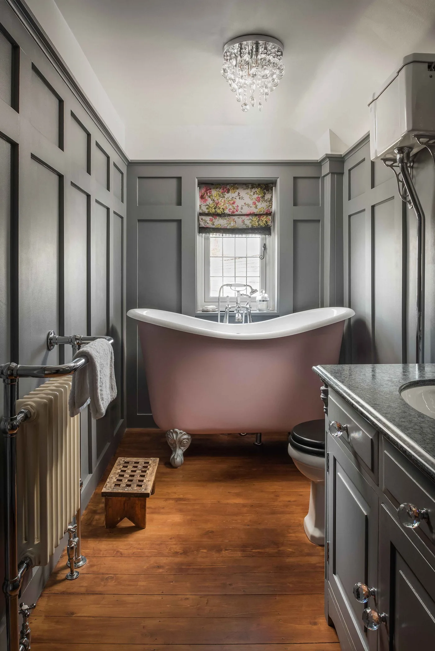 Narrow bathroom with gray wall paneling, a pink freestanding bath beneath a window, floral blind and gray basin cabinet.