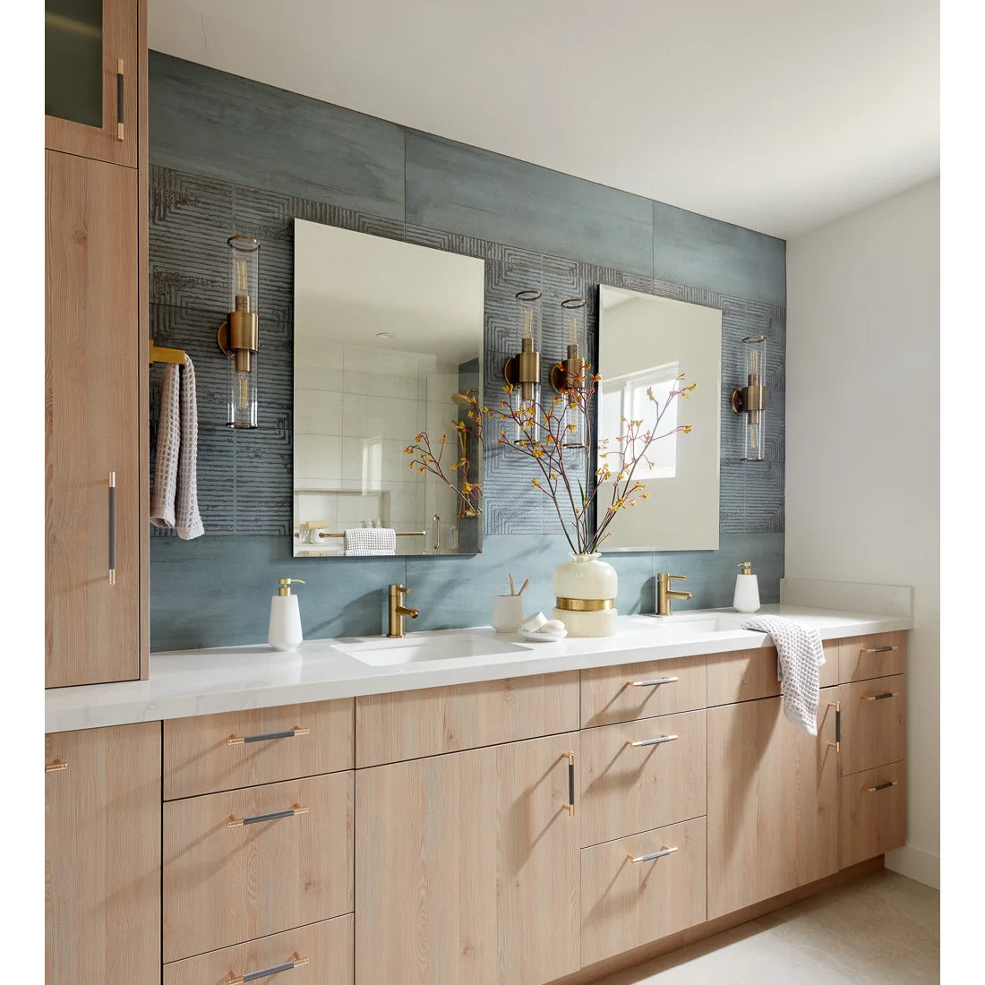 A pale wood double vanity stands beneath mirrors on a blue tiled wall with a darker textured band