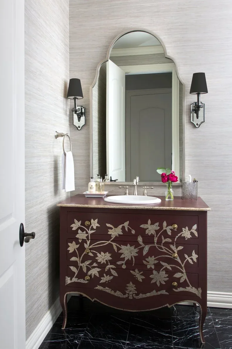 Powder room with curved dark wooden cabinet decorated with pale floral branches, white basin, shaped tall mirror, black-shaded wall lights and pale textured walls.