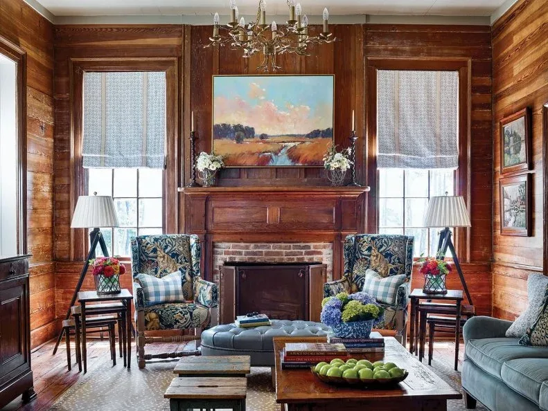 Brick fireplace with traditional wood mantel in a timber-paneled room, with landscape picture, patterned chairs and chandelier