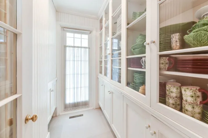 Narrow pale pantry with glazed dish cabinets, colorful crockery and tall window at the end