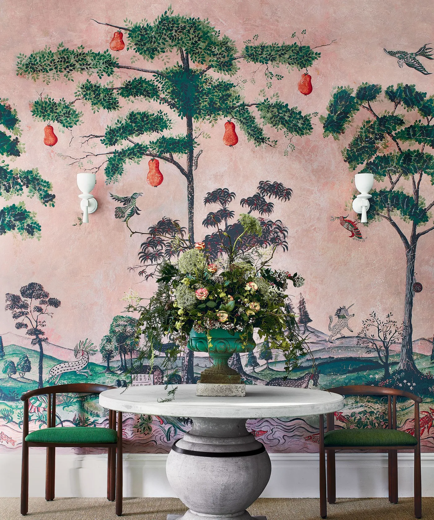 Pink landscape-mural wall behind a round pedestal table, green chairs and paired pale wall sconces.