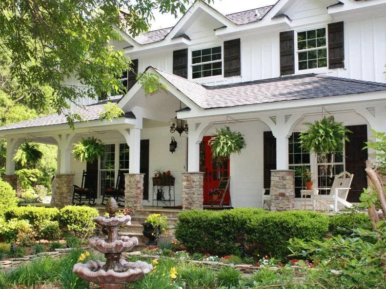 White country house with broad covered porch, hanging baskets, red door and mixed rocking chairs