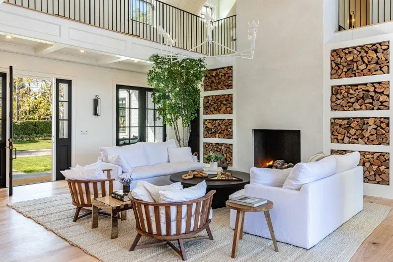 Tall plain pale fireplace with stacked-log shelf recesses on both sides, white sofas, wood chairs and an upper gallery