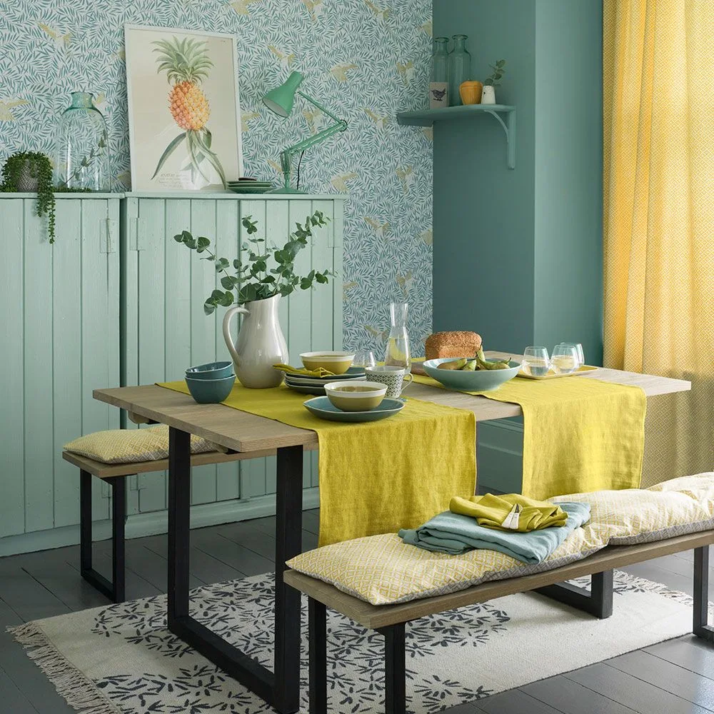 Yellow curtain beside a pale blue-green dining area with patterned wallpaper, a wooden table, yellow runners, cushioned benches, and a pineapple print.