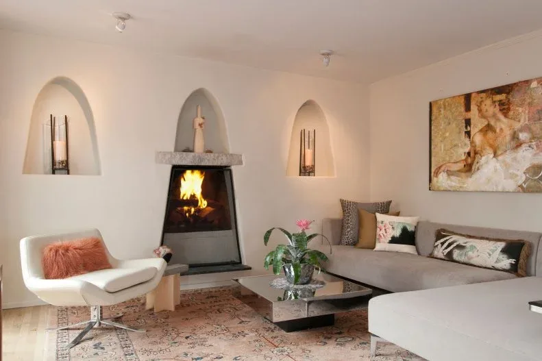 Neutral sitting room with pale walls and arched niches, dark fireplace, gray sectional, white curved chair and patterned pale rug.