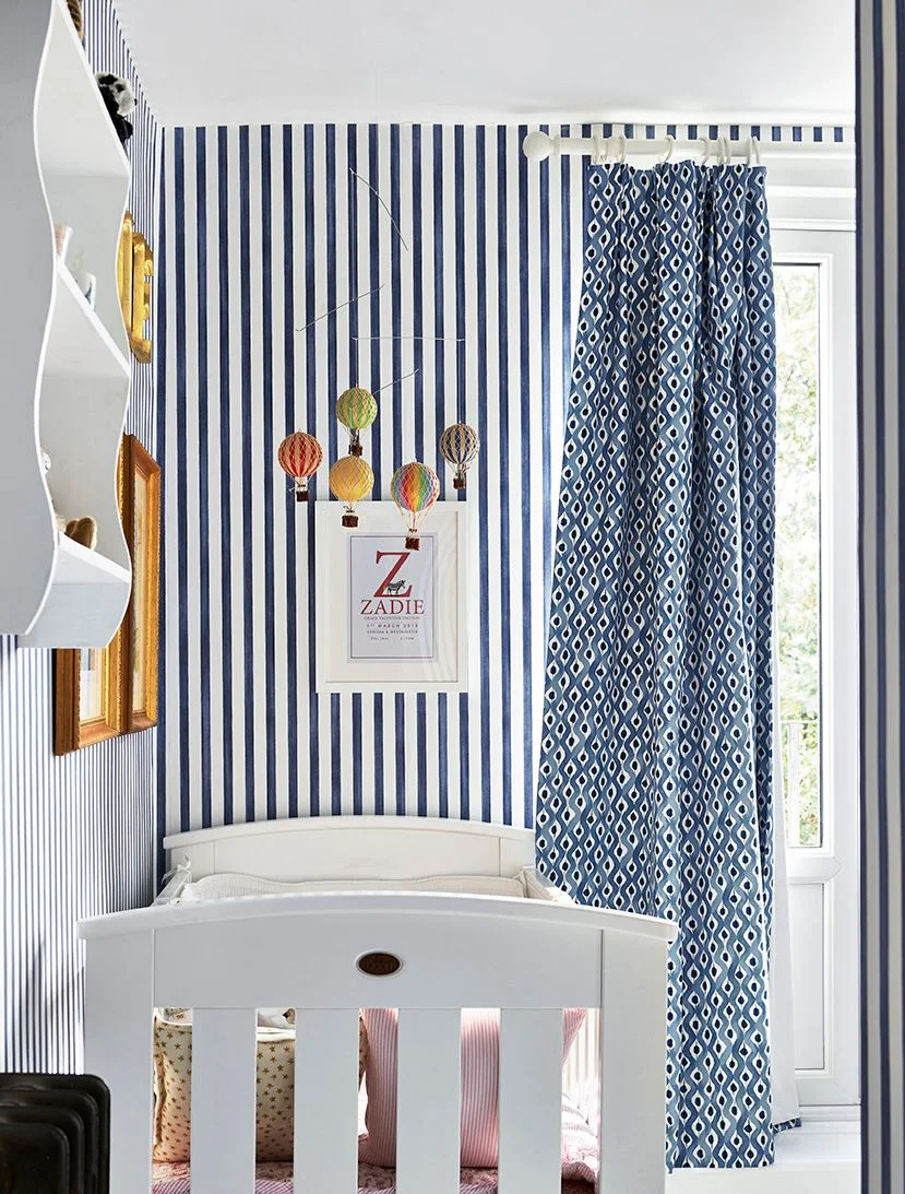 Navy-and-white vertical nursery stripes surround a white crib beside blue patterned curtains, framed art and colorful hanging balloon decorations
