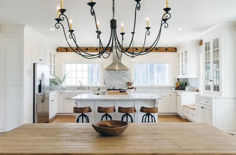 White kitchen cabinets stand behind a broad timber dining table and dark chandelier