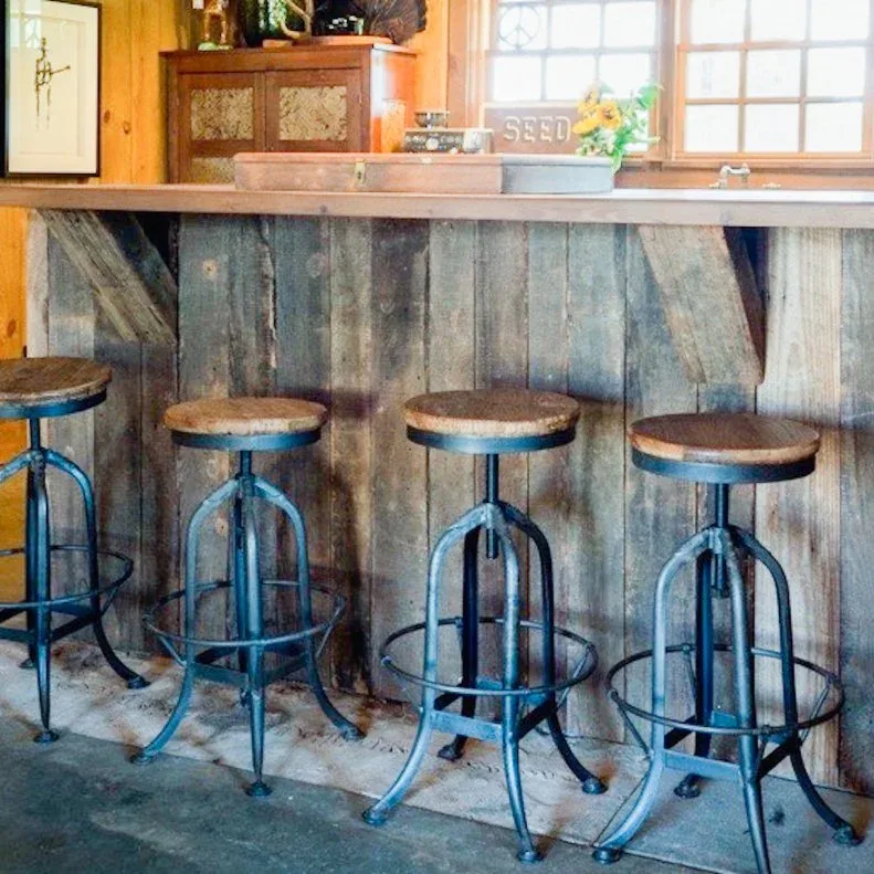 A weathered-looking wood breakfast bar with diagonal board details sits beside blue metal-framed stools below warm wooden walls and a sink window