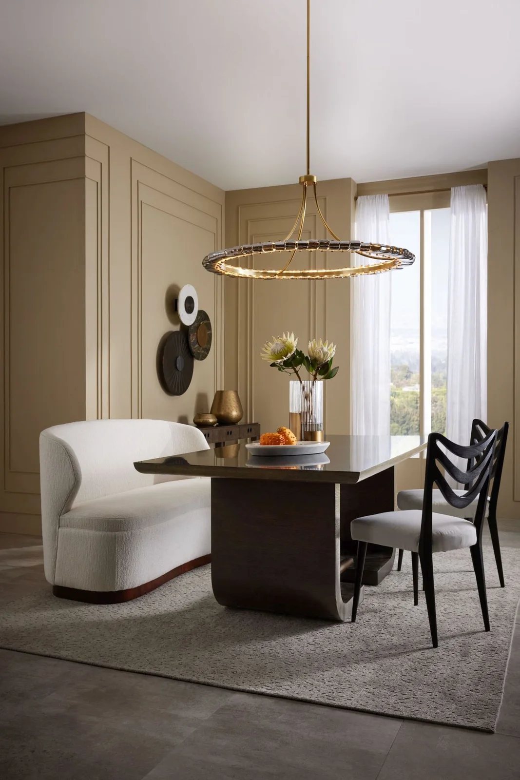 A pale curved dining bench sits beside a dark rectangular table and black chairs below a ring pendant against muted paneled walls and light curtains