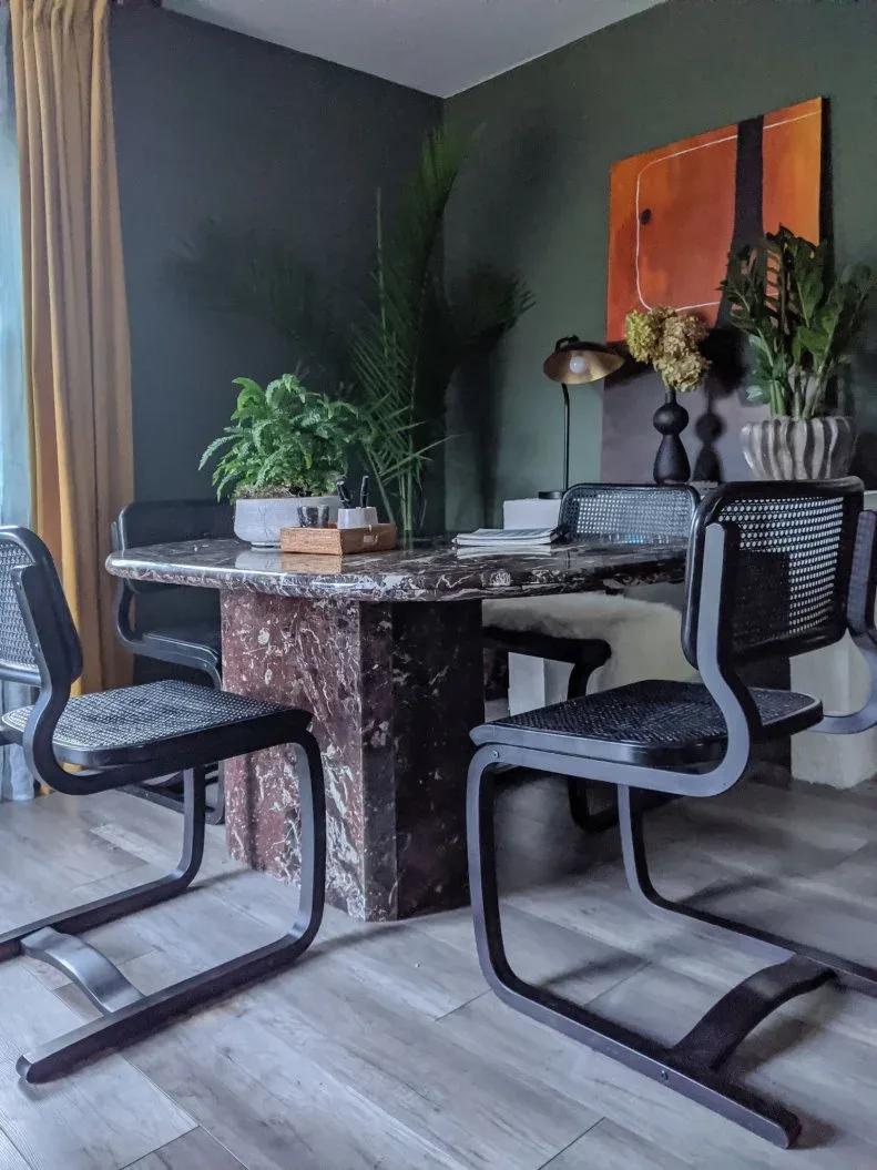 Dark veined dining table with black mesh-backed chairs, plants and a desk lamp against dark walls