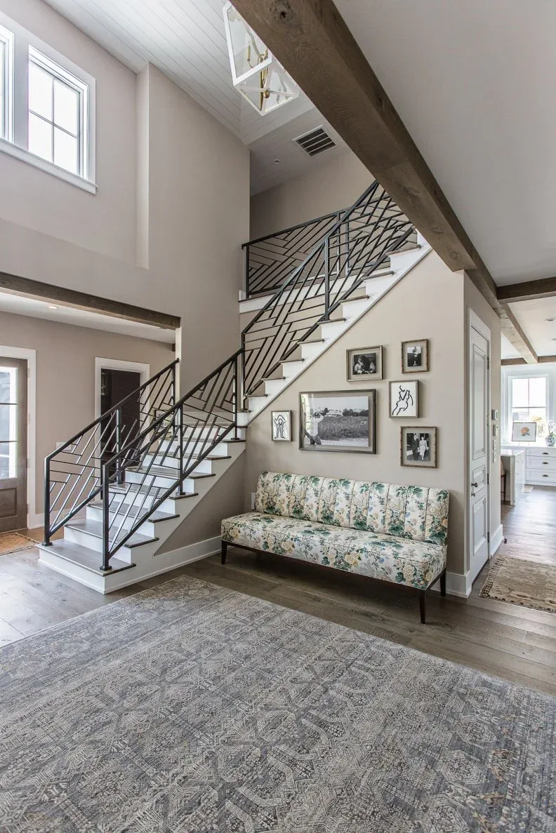 Pale entrance with floral upholstered settee beneath staircase, dark geometric railing, exposed beam and large light patterned rug.