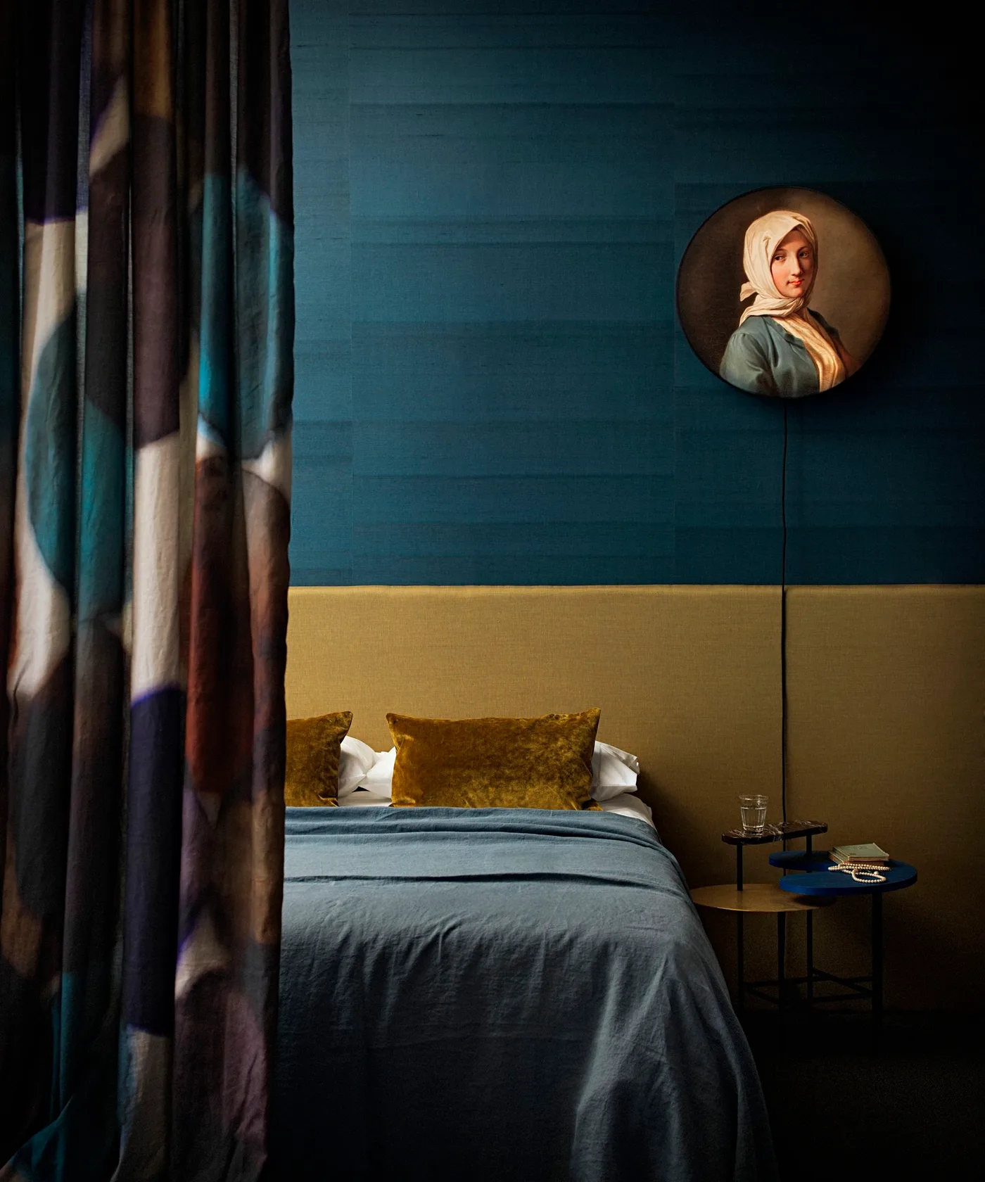 A warm ochre-colored headboard and cushions sit against a deep teal paneled wall beside a small round table and tall portrait light