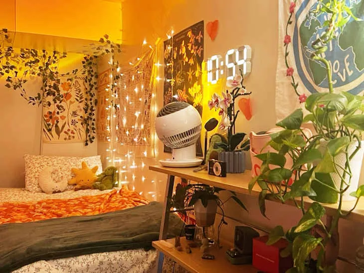 Warmly lit dorm room with trailing greenery, string lights, floral bedding and open shelving beside the bed.