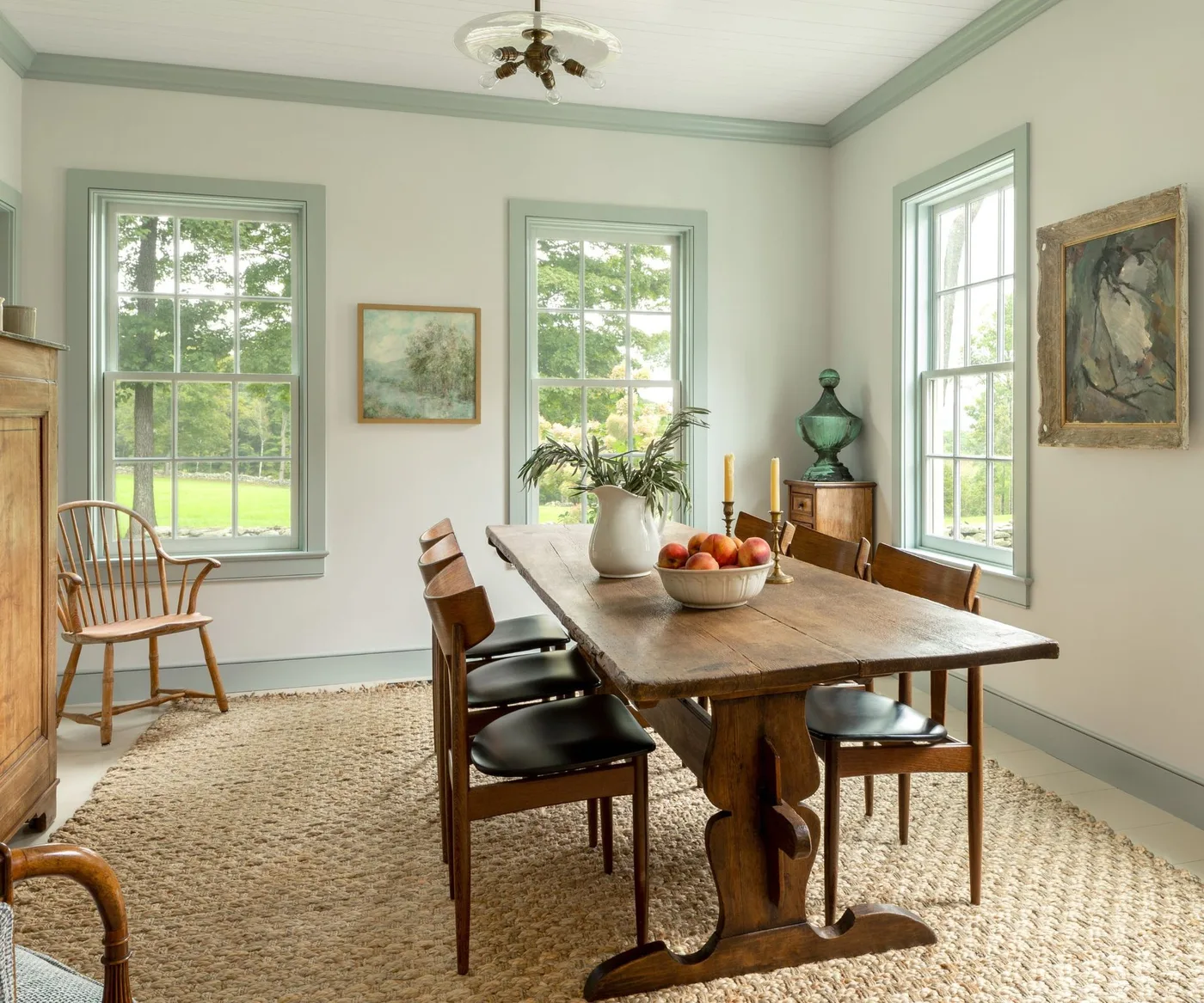 Long wooden trestle table with dark-seated chairs, pale green window trim and a textured neutral rug.