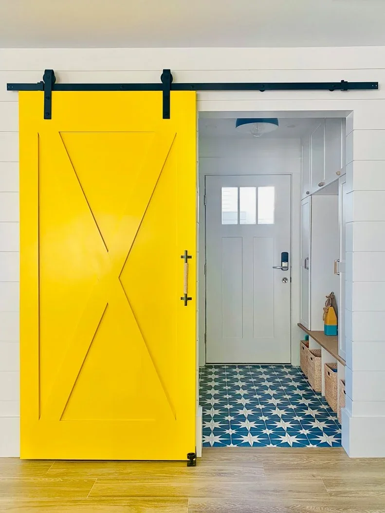 A yellow sliding door with crossed panels stands beside white horizontal boards, opening onto pale coat storage, baskets and a blue patterned entry floor