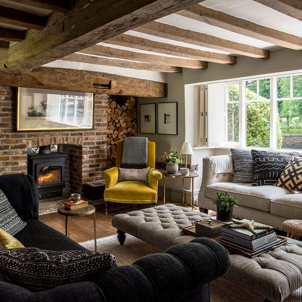 Pale seating, a yellow armchair and tufted ottoman sit beneath exposed wooden beams beside a brick fireplace