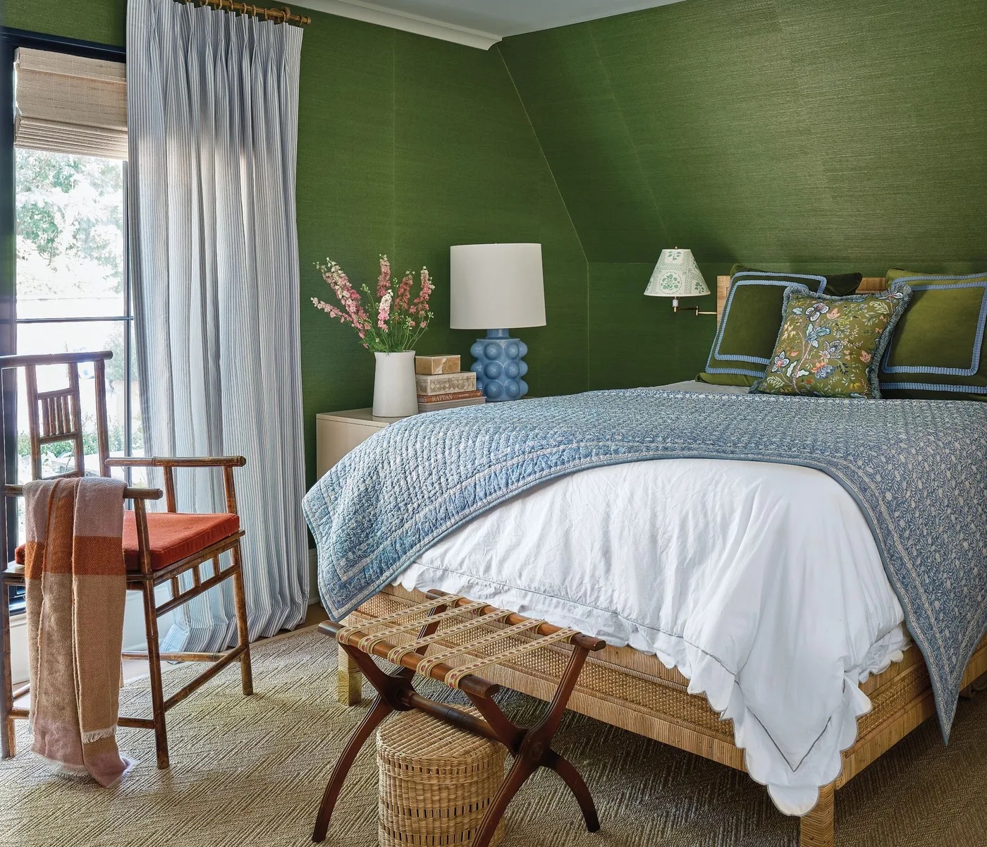 Green bedroom with pale blue curtains, blue patterned bedding, a woven headboard and a wooden chair with a warm-colored seat.