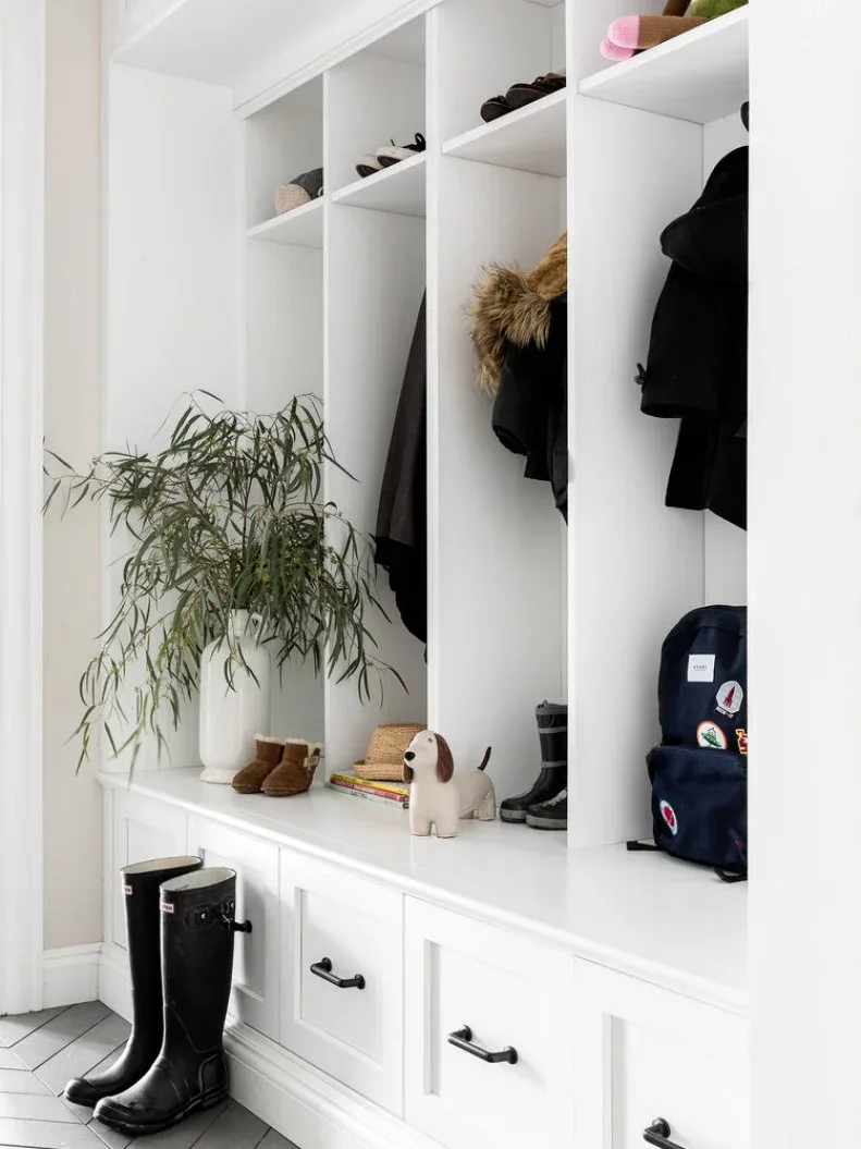 White open coat compartments and shoe shelves stand above framed bench drawers with dark handles beside pale walls and a pale floor