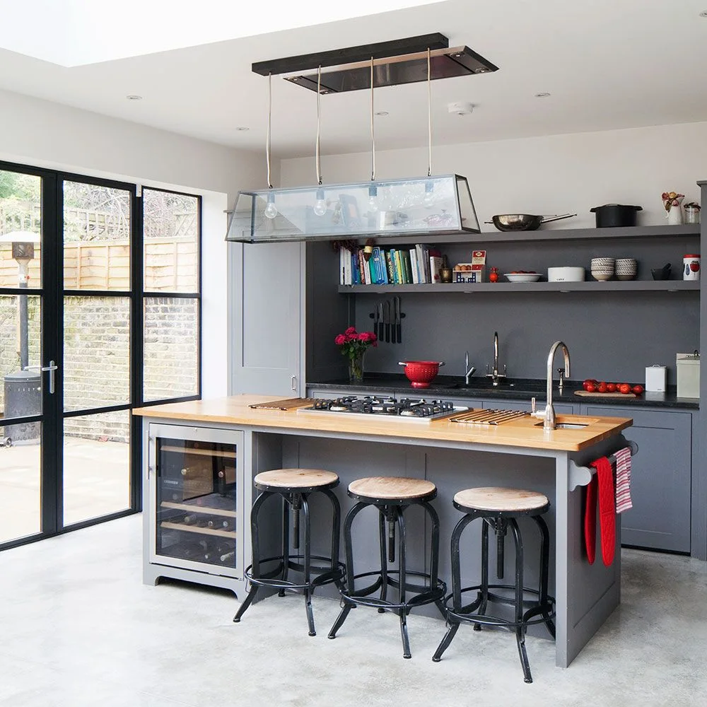 A gray island with a wooden counter, sink, cooktop and bottle cooler sits before dark gray shelves beside three round stools