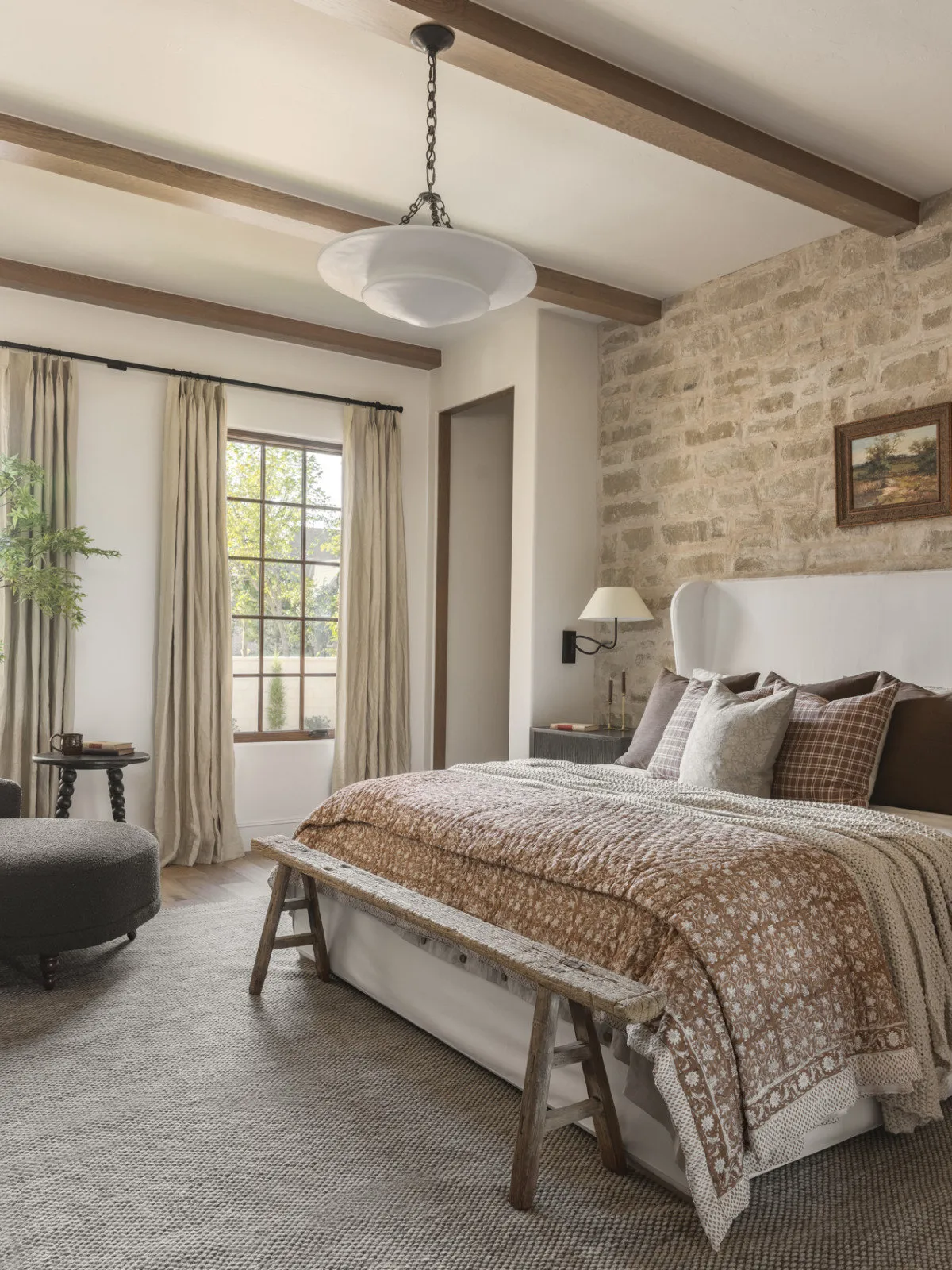 A pale bed stands against textured stonework beneath timber beams, with light furnishings nearby.