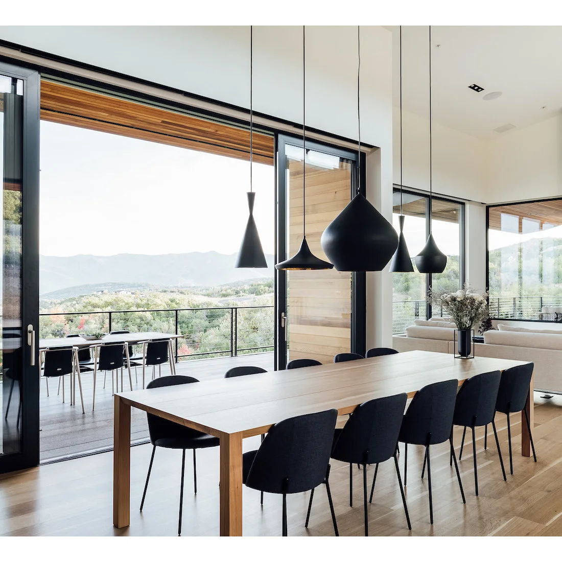 A long pale wood dining table and dark slim chairs stand beneath five differently shaped dark pendants beside black-framed glazing and outdoor seating