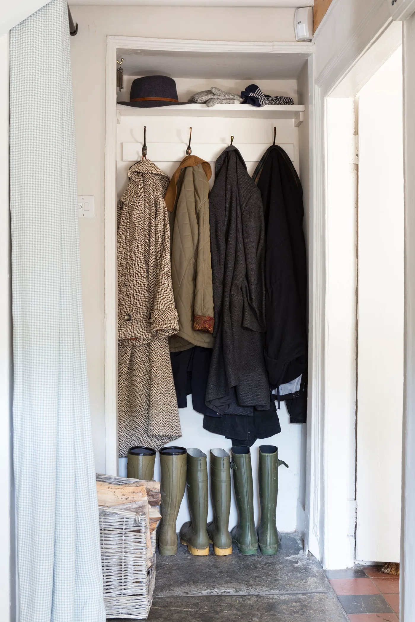 Narrow white entry recess with four hanging coats, an upper hat shelf, tall boots and a curtain at one side