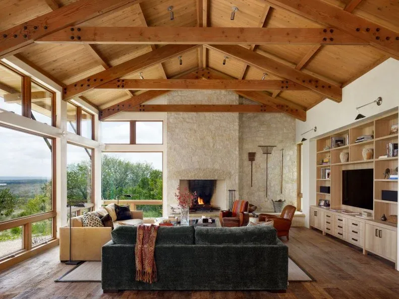 Tall pale textured fireplace at the end of a timber-trussed great room with wood ceiling, dark sofa and open shelving