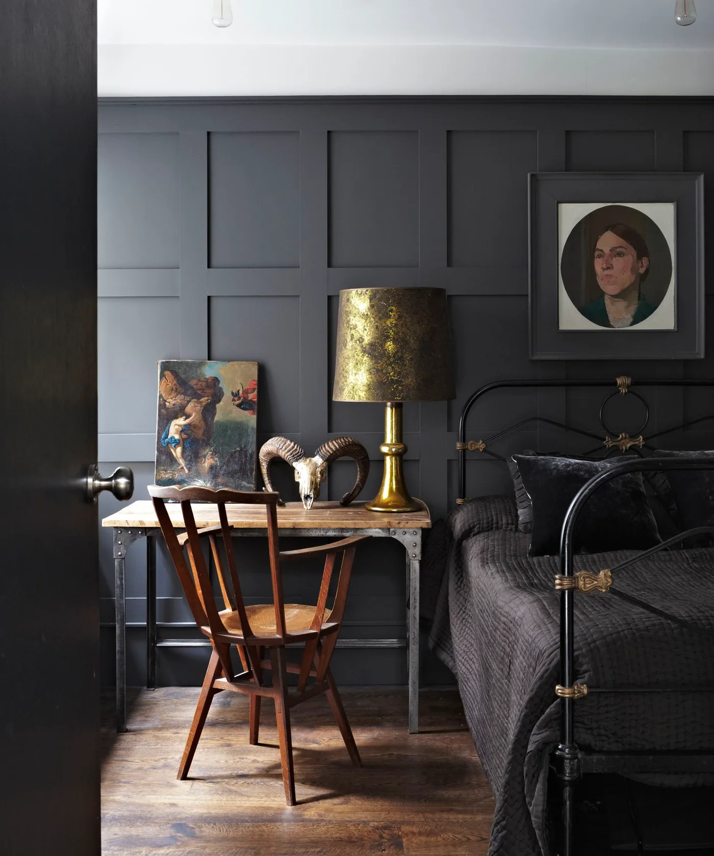 A dark paneled bedroom has an open dark bed frame beside a wooden desk and chair with a warm-colored table lamp and framed portrait