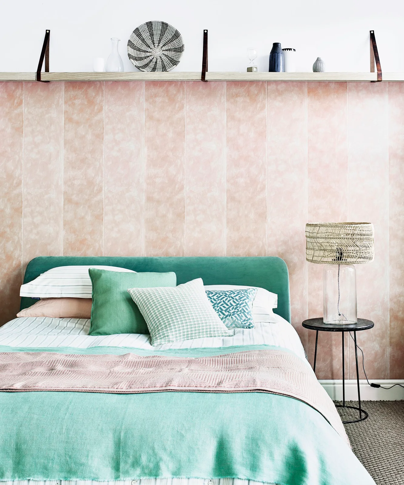 A green upholstered headboard and pale green throw sit beside white bedding against a softly patterned pink wall with a long shelf