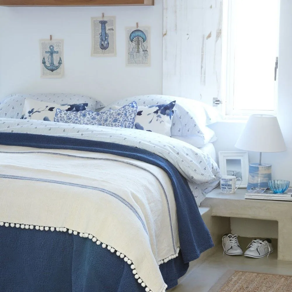 Bright white bedroom with blue-and-white patterned bedding, small nautical wall pictures and a pale bedside table with open storage.