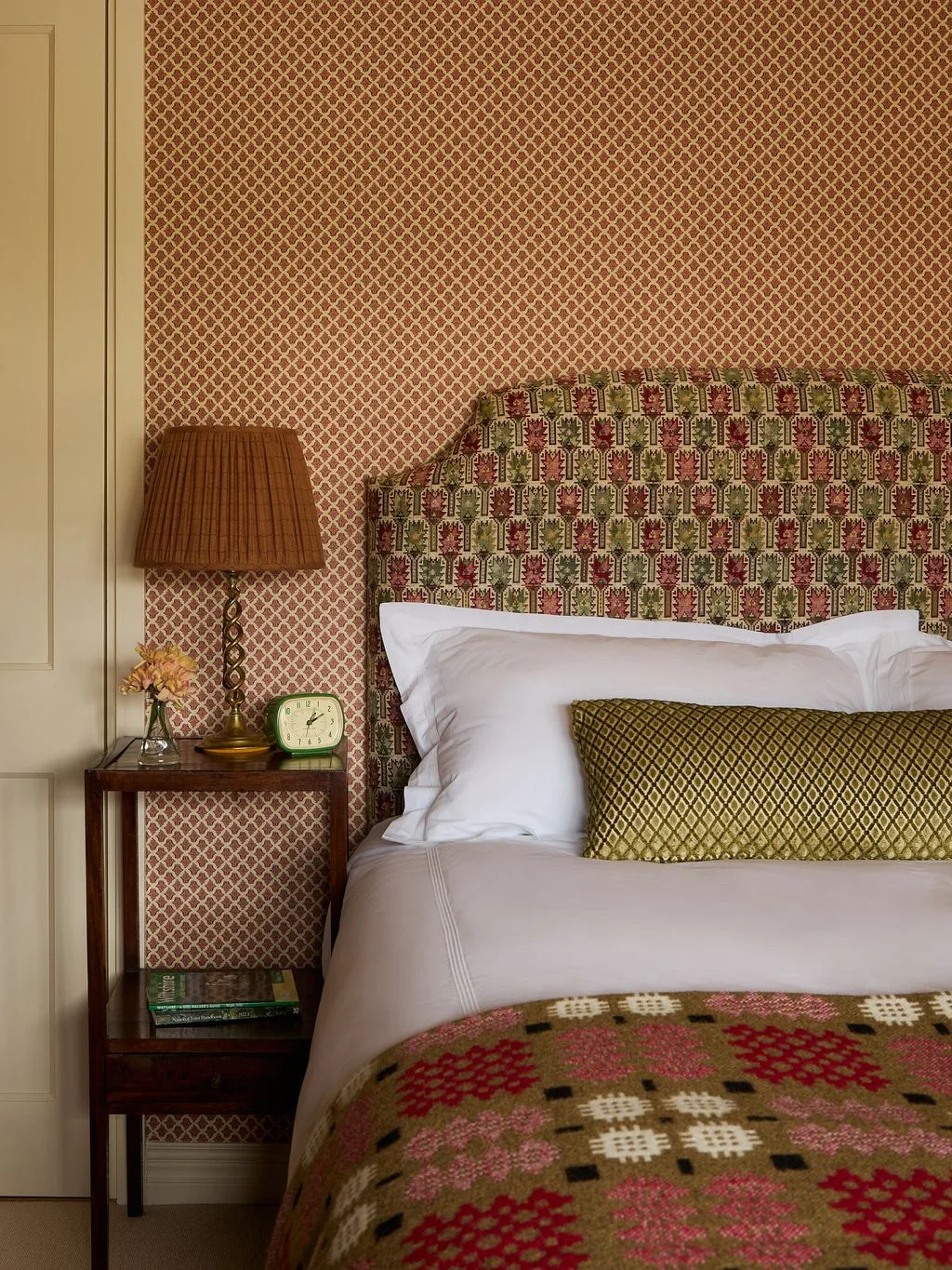 Bedroom with fine reddish geometric wallpaper, shaped patterned headboard, white pillows, olive cushion and colorful geometric bed cover