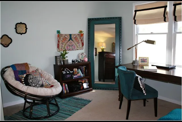 A round woven chair sits beside a dark desk in a bedroom with colorful pillows.