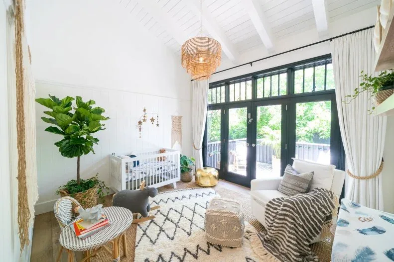 A pale armchair, white crib and small woven chairs stand in a bright nursery with a high ceiling and black-framed doors