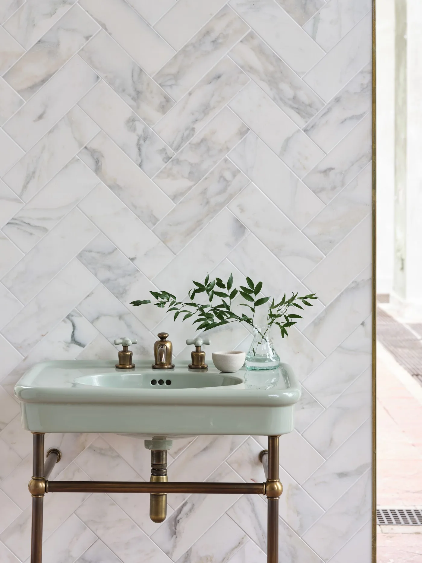 Pale green wash basin on open warm-toned metal frame against pale herringbone-pattern wall