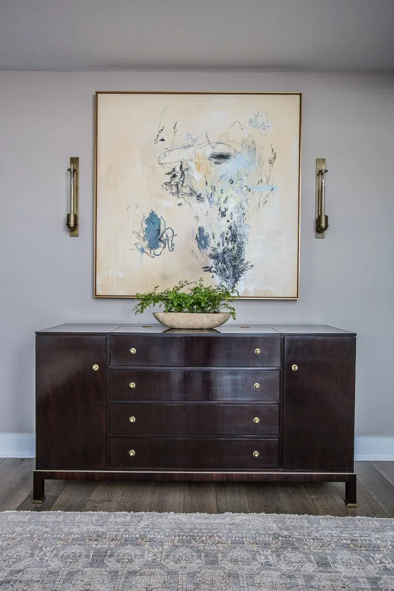 Gray hall with dark wooden drawer cabinet, large pale abstract artwork, paired slender sconces and shallow plant bowl.