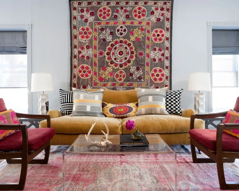 Living room with mustard-yellow sofa, pink-upholstered wooden chairs, pink rug, colorful textile wall hanging, black-and-white cushions and clear table.