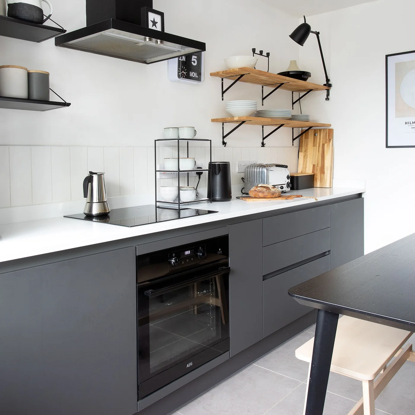 Charcoal gray flat cabinets and drawers run below a white countertop and tiled wall beside an oven and open wooden shelves