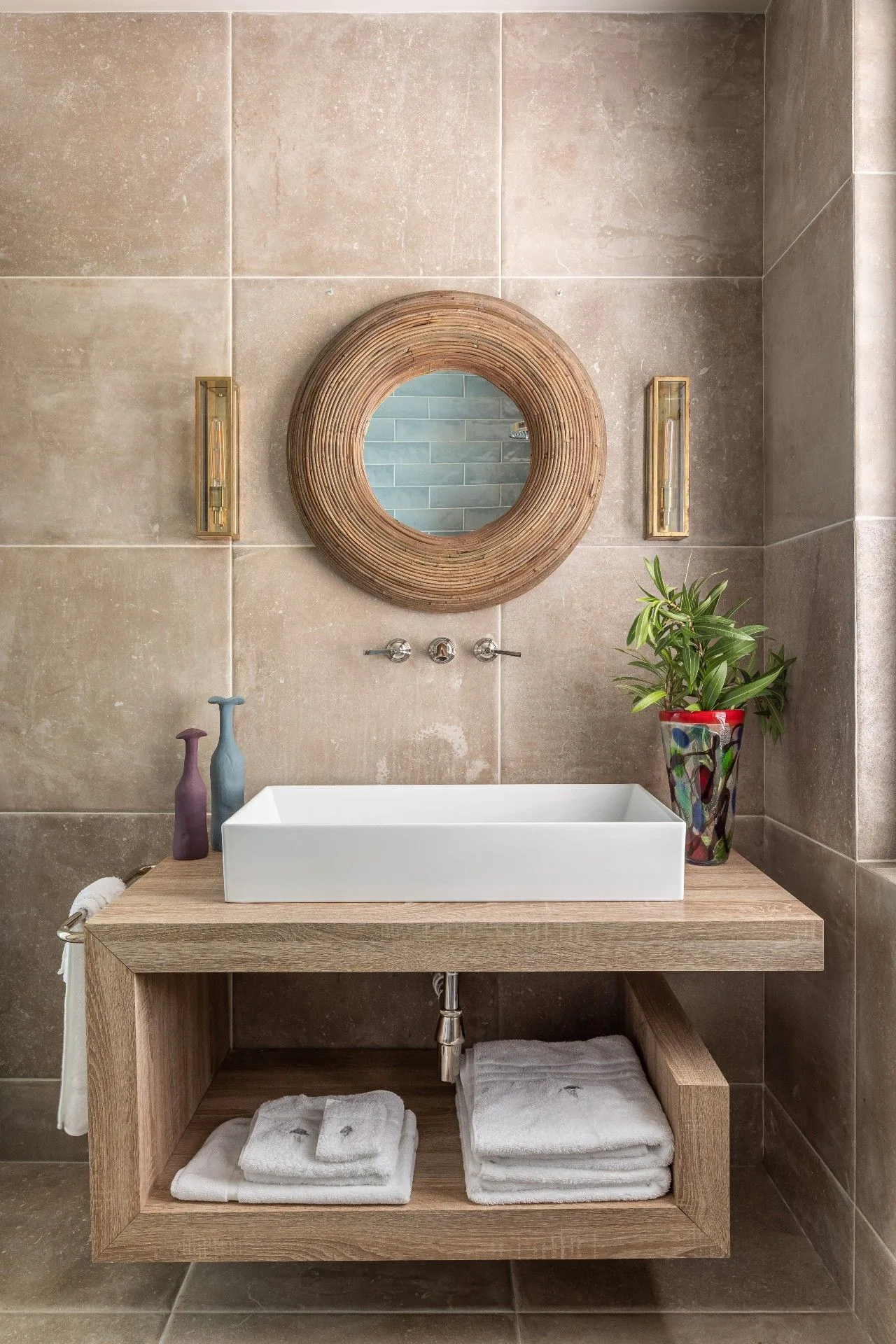 Powder room with round woven mirror, beige tiled walls, rectangular white basin on wooden console, folded towels and a leafy plant in colorful container.