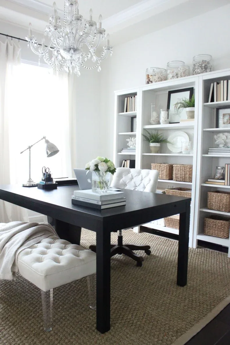 Dark rectangular work table with a laptop, white tufted chairs, bookshelves and a chandelier