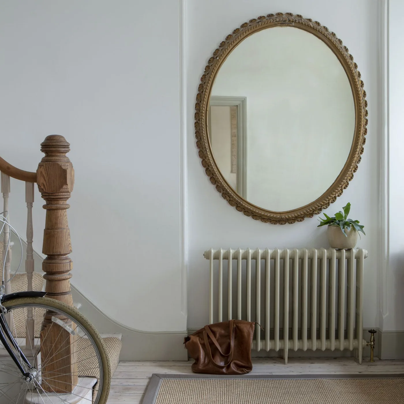 White entry corner with large oval gold-colored framed mirror over pale radiator, wood stair newel, bicycle and neutral floor rug