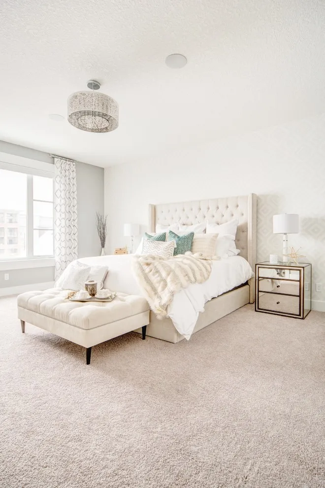 A pale bed and upholstered ottoman sit beside a mirrored chest in a bright room