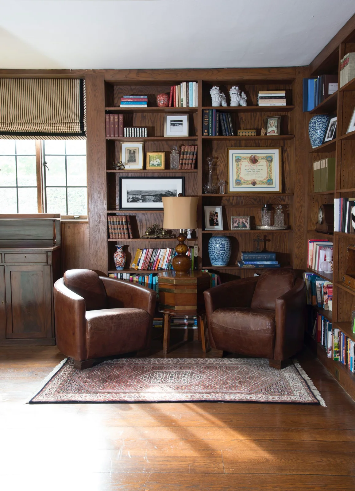 Wooden bookshelves turn a corner behind two brown chairs, a small shared table and a patterned rug near a window