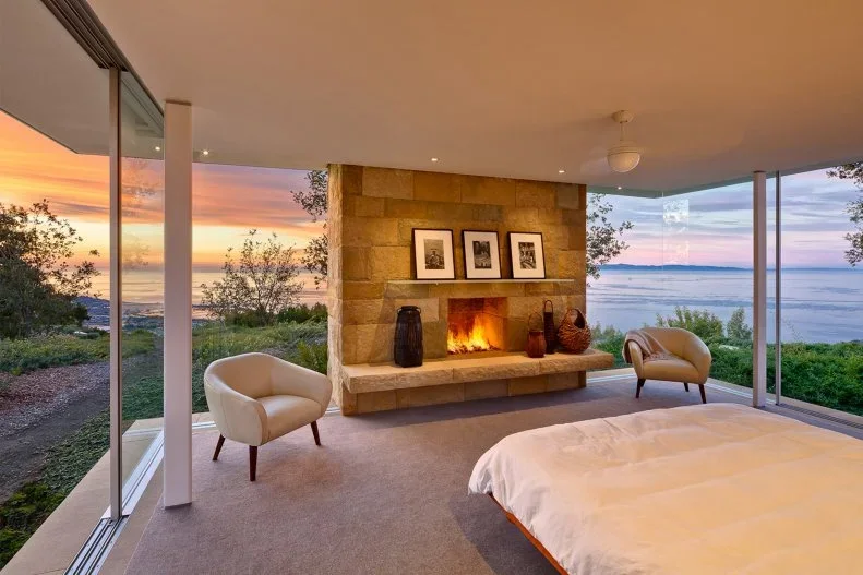 A bedroom fireplace wall with broad warm-colored blocks and a long hearth stands between glazed walls beside two pale chairs and a bed