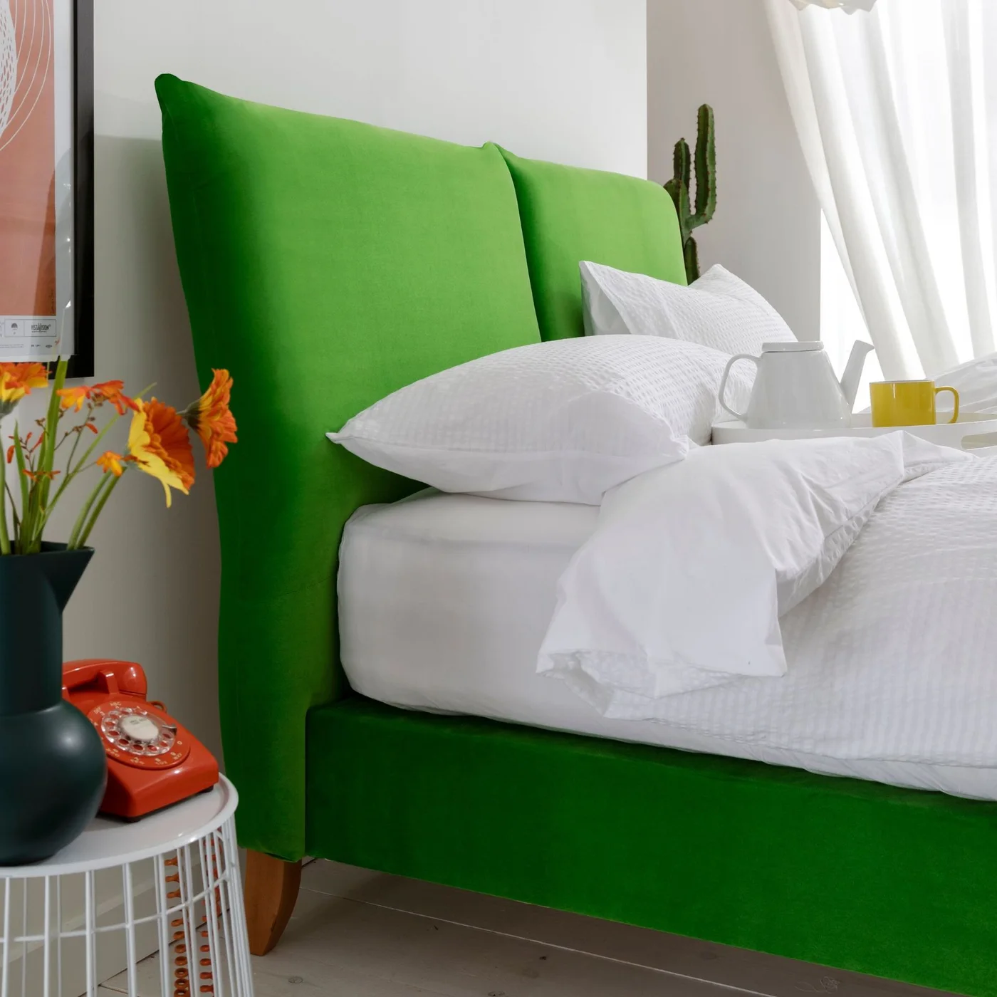 A bright green upholstered bed holds white pillows and bedding beside a pale round table, orange phone and yellow mug