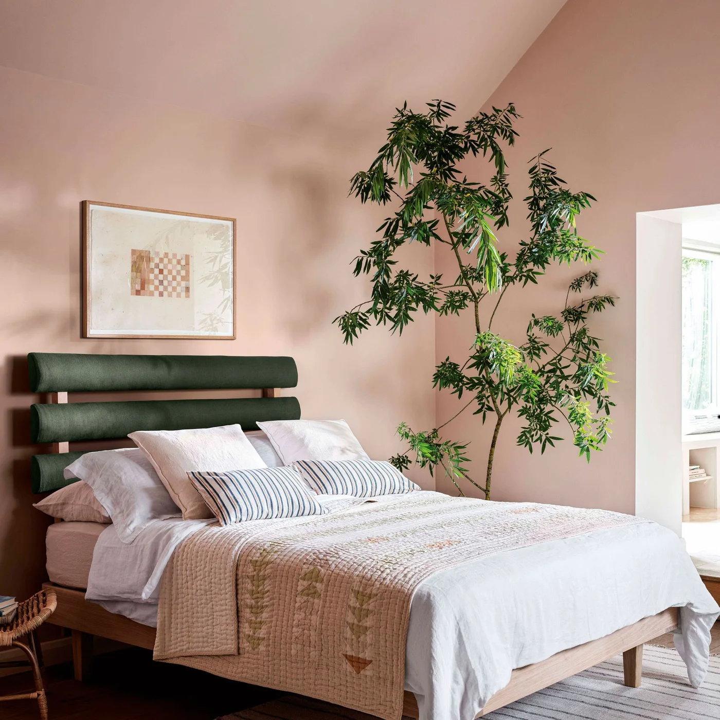 A wooden bed with a two-part dark green headboard sits against pale pink walls beside striped cushions, framed art and a tall leafy plant