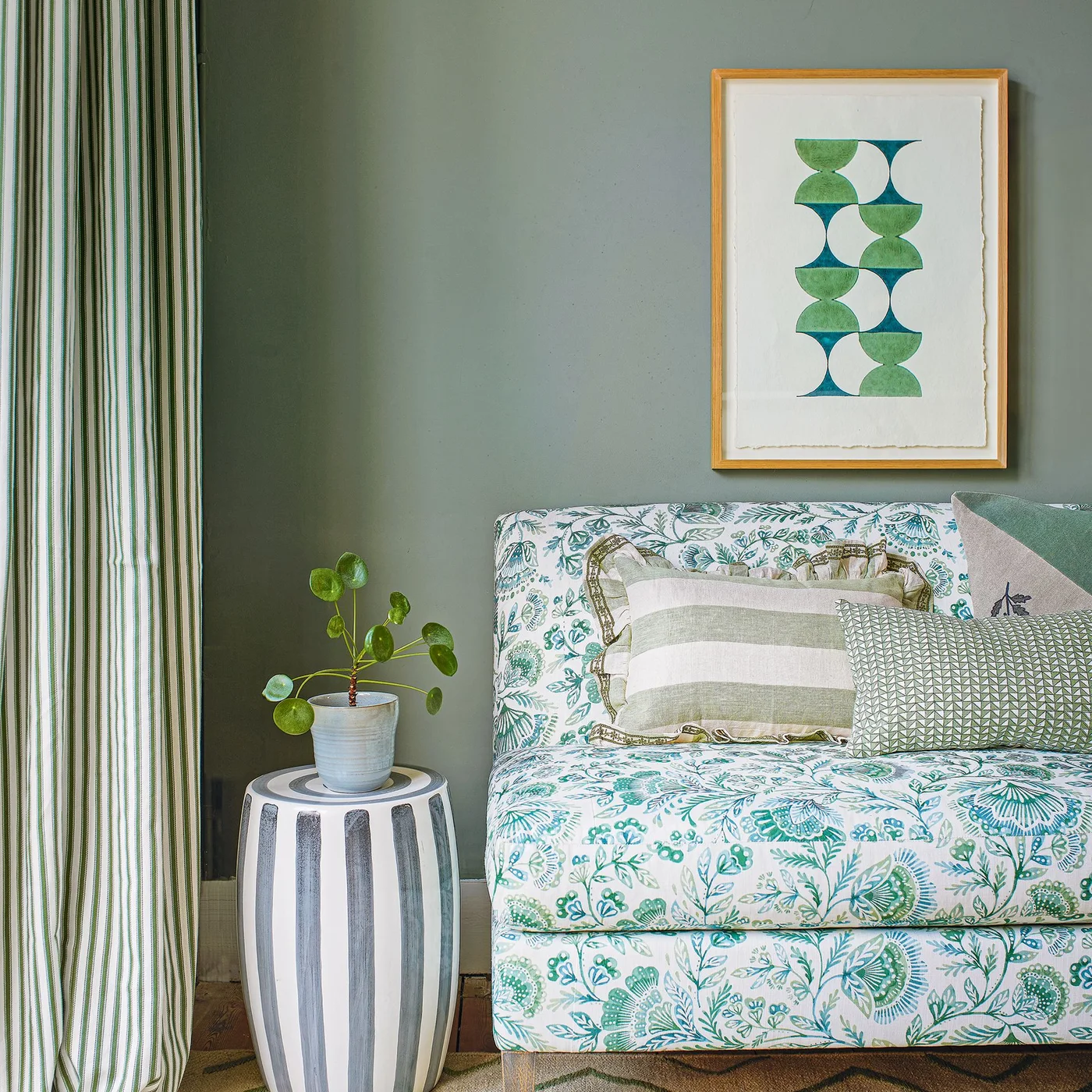 A pale sofa with green floral upholstery sits beside striped curtains and a striped round side table against gray-green walls